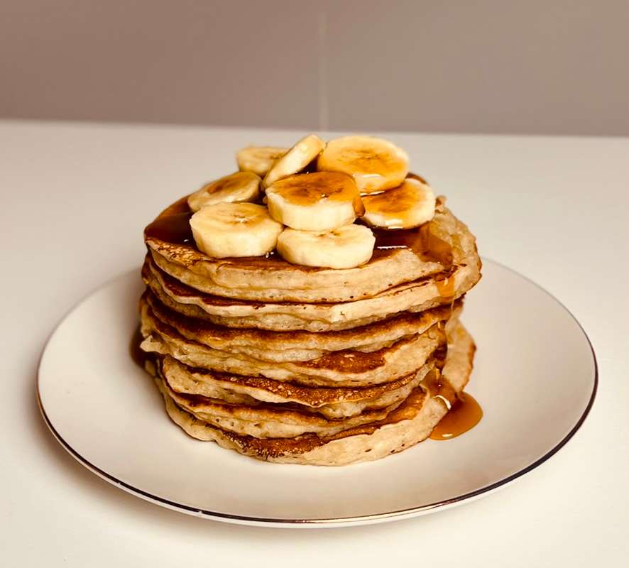Stacked banana pancakes on a white plate. The pancakes are topped with sliced bananas and a drizzle of syrup.
