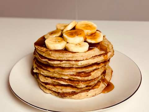 Stacked banana pancakes on a white plate. The pancakes are topped with sliced bananas and a drizzle of syrup.