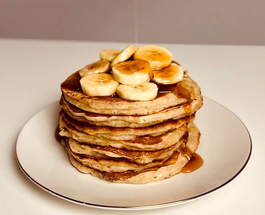 Stacked banana pancakes on a white plate. The pancakes are topped with sliced bananas and a drizzle of syrup.