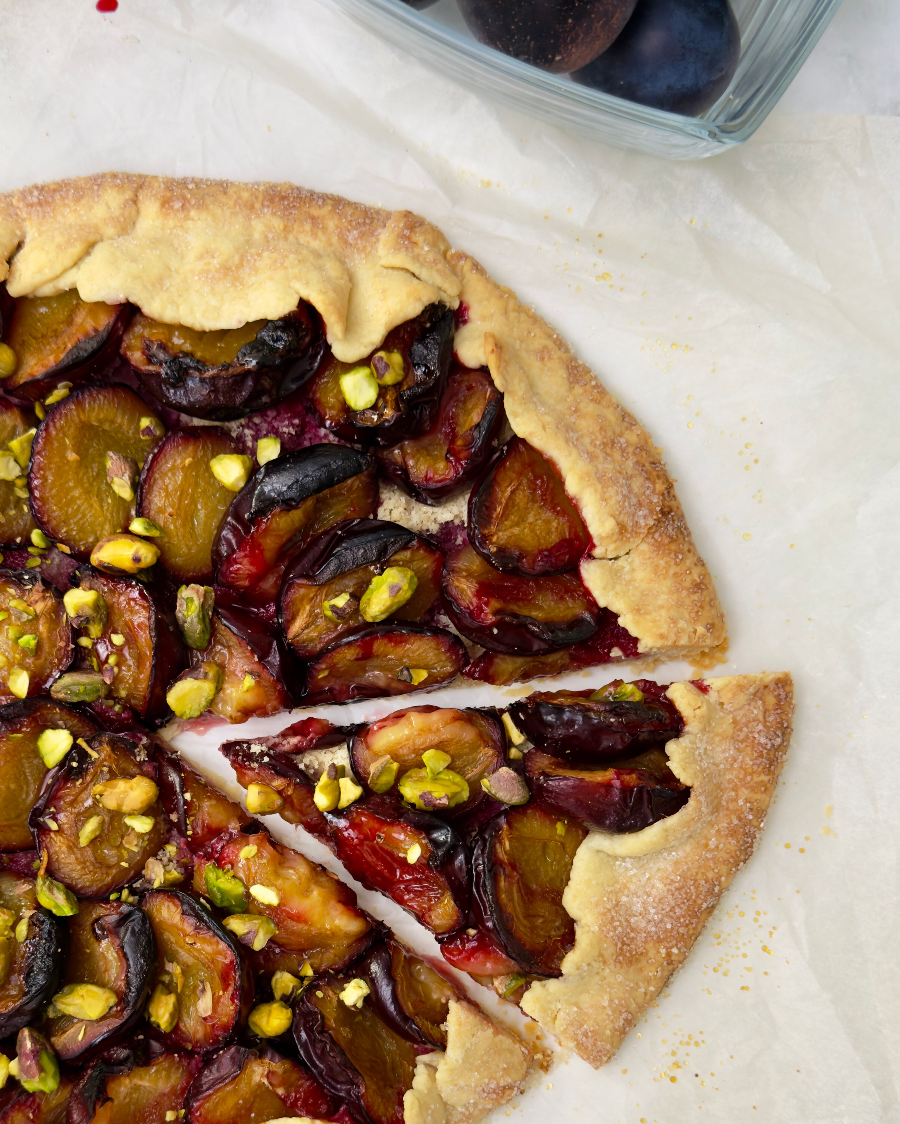 Close-up of a partially sliced, rustic plum galette on parchment paper. The galette has a golden, flaky crust sprinkled with sugar and is filled with halved, deep purple plums and green chopped pistachios.