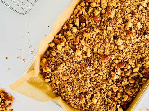 Overhead close-up of crunchy granola clusters with visible peanuts and oats spread on a baking sheet.
