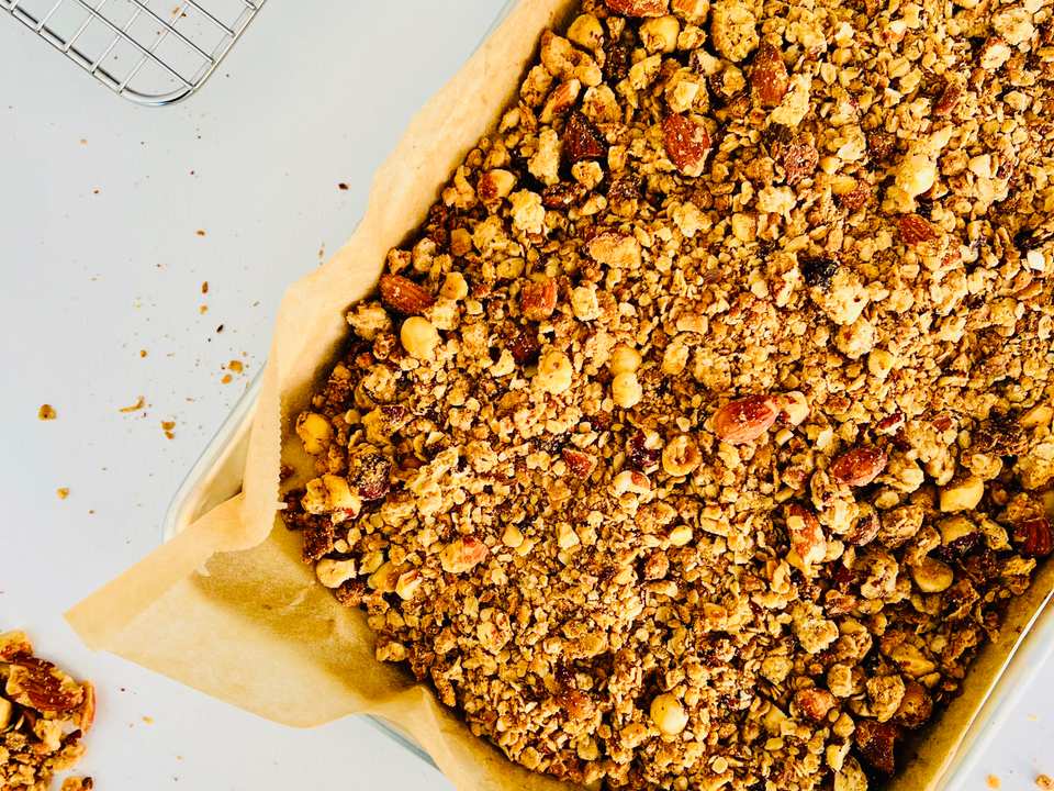 Overhead close-up of crunchy granola clusters with visible peanuts and oats spread on a baking sheet.
