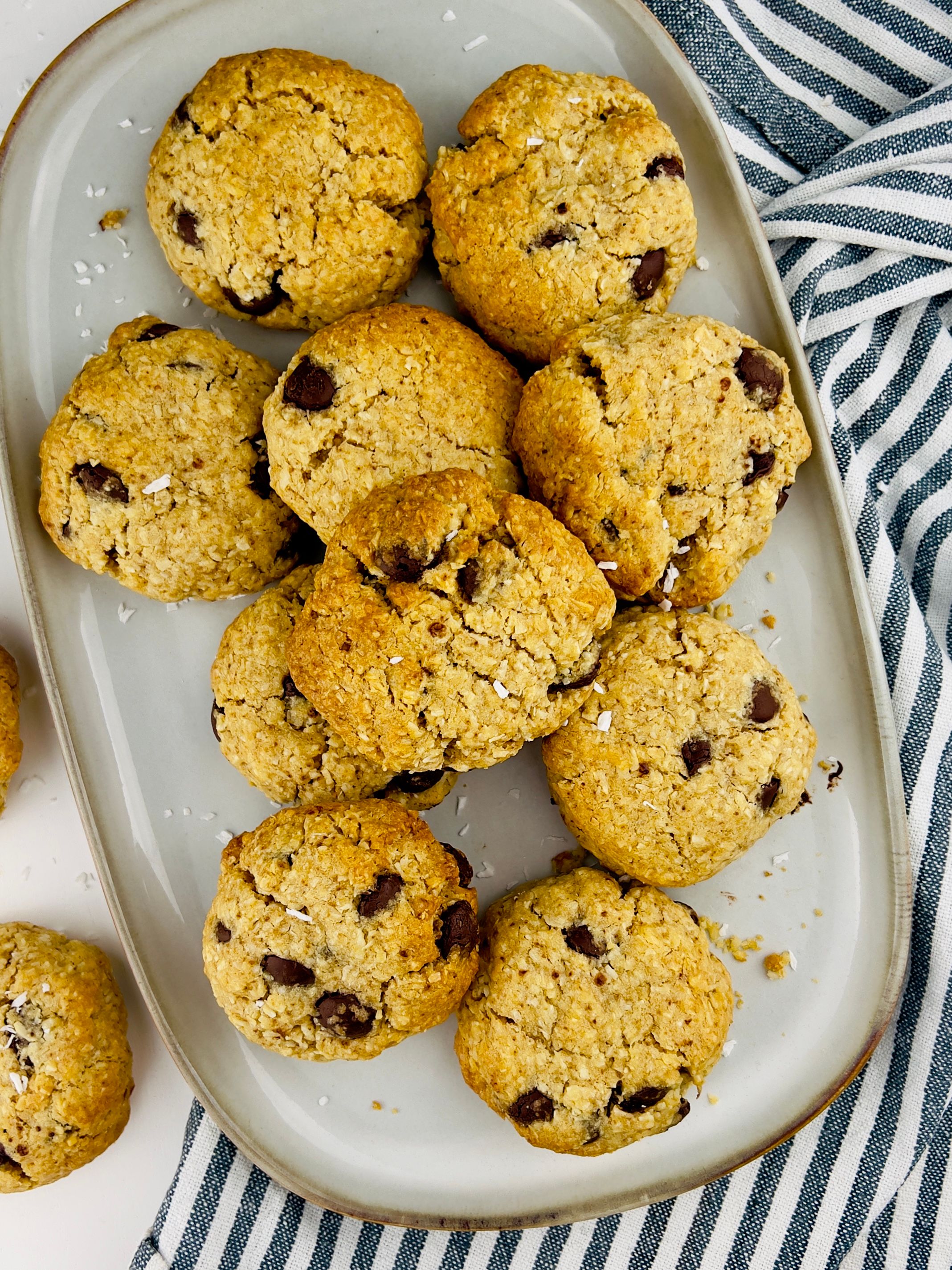 Overhead shot of many round coconut-oatmeal chocolate chip cookies with flakes of coconut on a gray dish, with a striped cloth on the side.