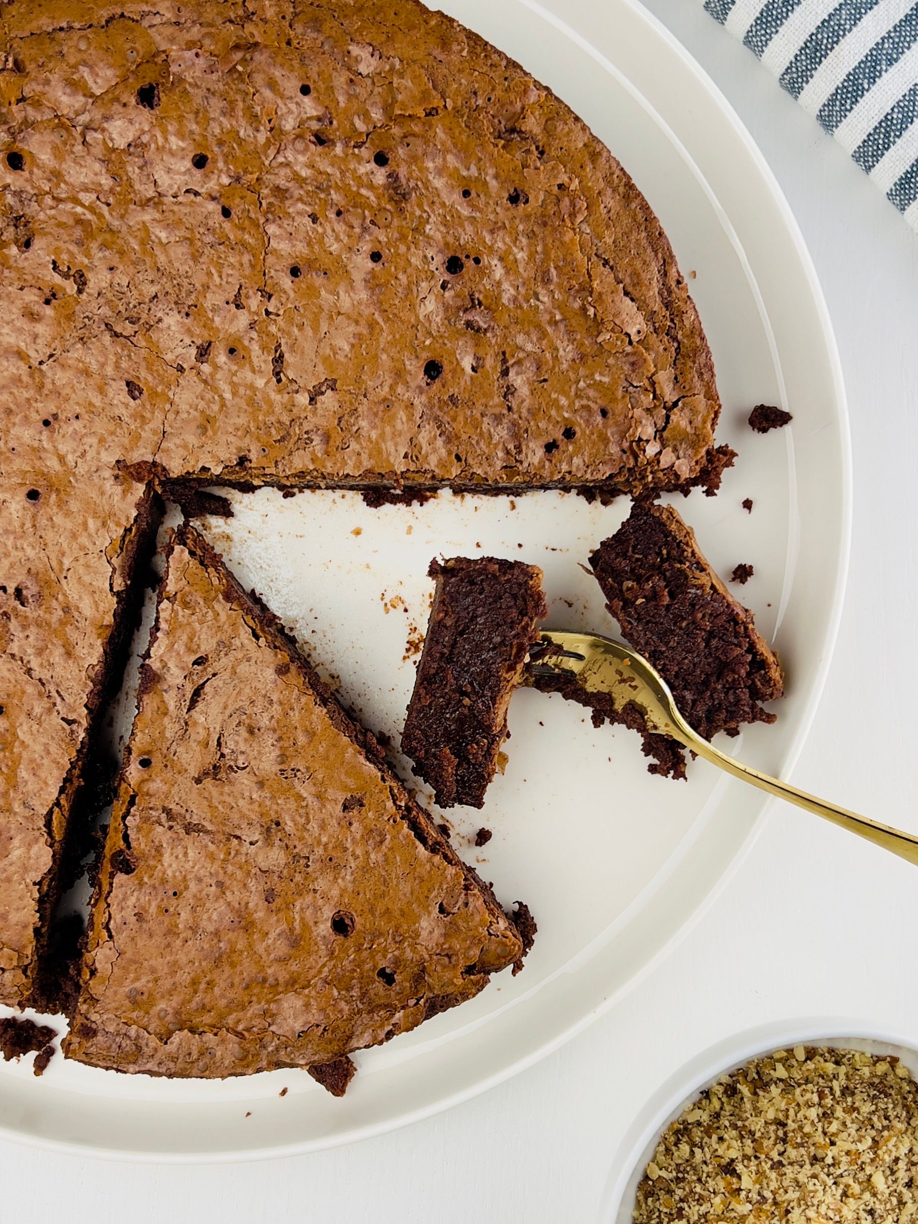A round, dark chocolate cake with a cracked top and visible hazelnuts, dusted lightly, on a cooling rack.
