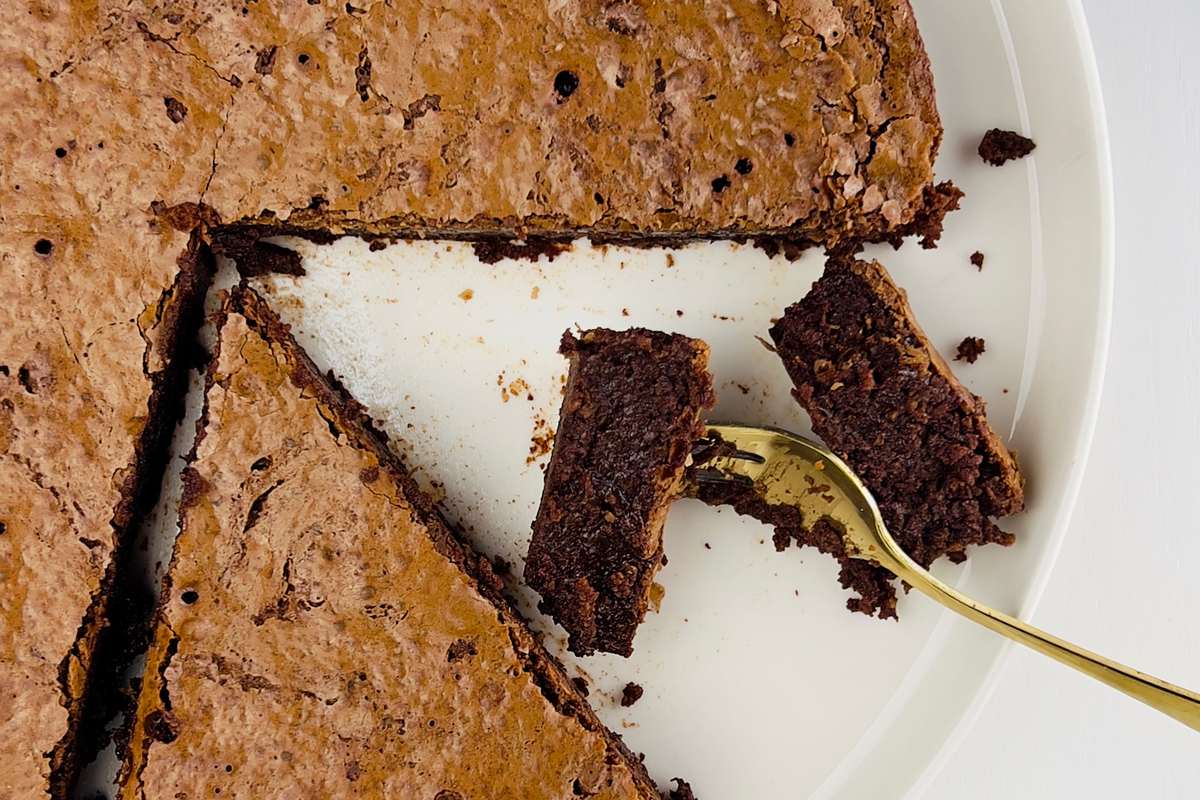 A round, dark chocolate cake with a cracked top and visible hazelnuts, dusted lightly, on a cooling rack.