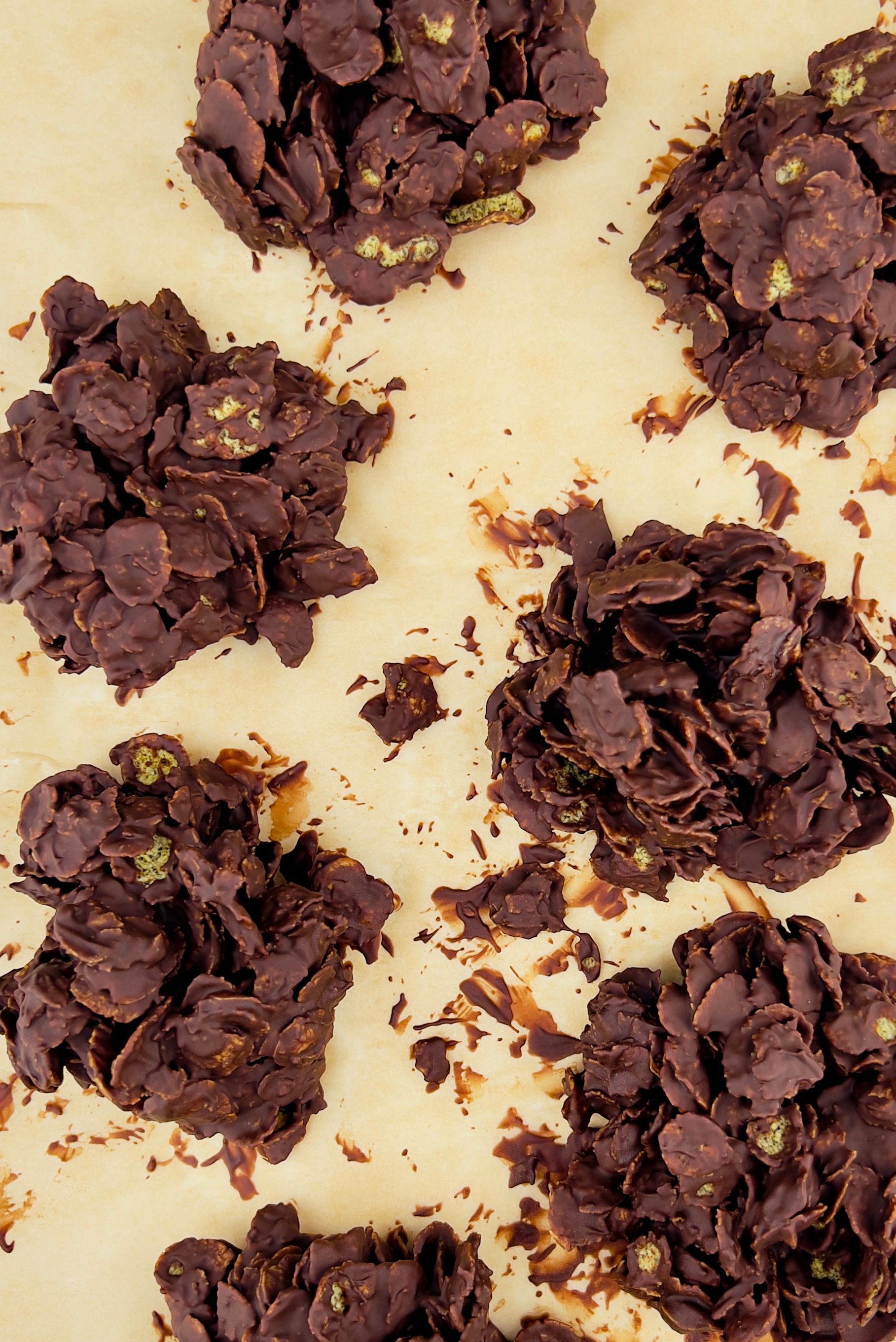 Overhead view of five clumps of chocolate cornflake clusters with pistachio pieces on parchment paper.