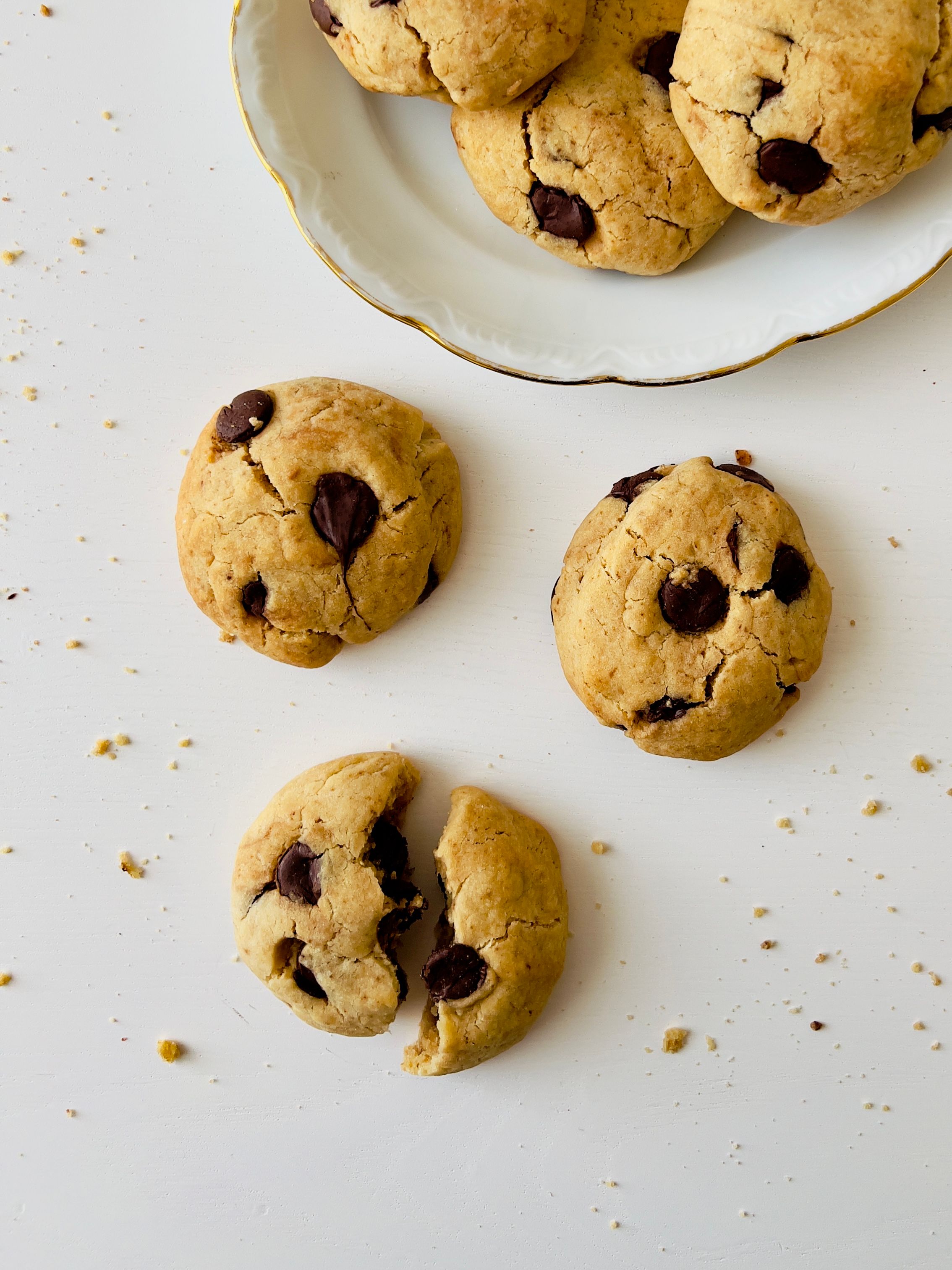 Overhead view of a white, gold-rimmed plate of chocolate chip cookies with 3 cookies scattered on the white table. One cookie is broken in half, revealing melted chocolate chips.