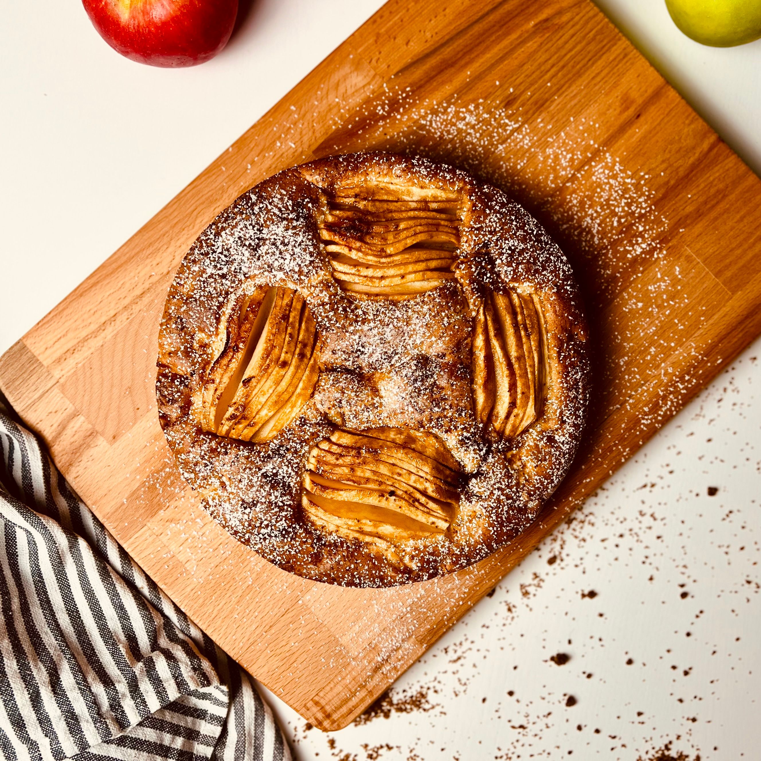 Overhead view of a round golden-brown apple cake dusted with powdered sugar on a wooden cutting board, with a striped linen and red and green apples.