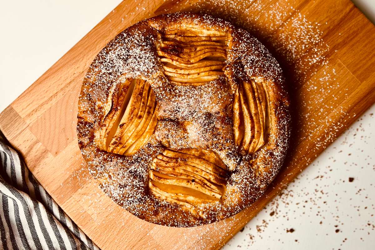Overhead view of a round golden-brown apple cake dusted with powdered sugar on a wooden cutting board, with a striped linen and red and green apples.