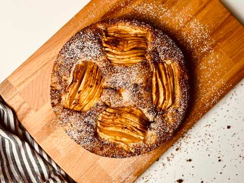 Overhead view of a round golden-brown apple cake dusted with powdered sugar on a wooden cutting board, with a striped linen and red and green apples.