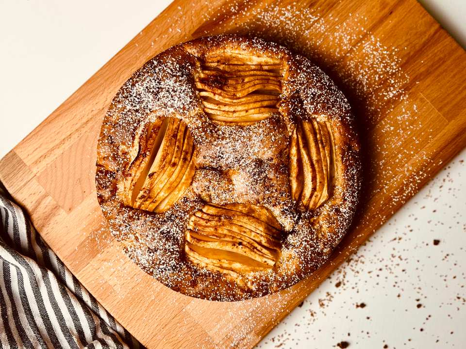 Overhead view of a round golden-brown apple cake dusted with powdered sugar on a wooden cutting board, with a striped linen and red and green apples.