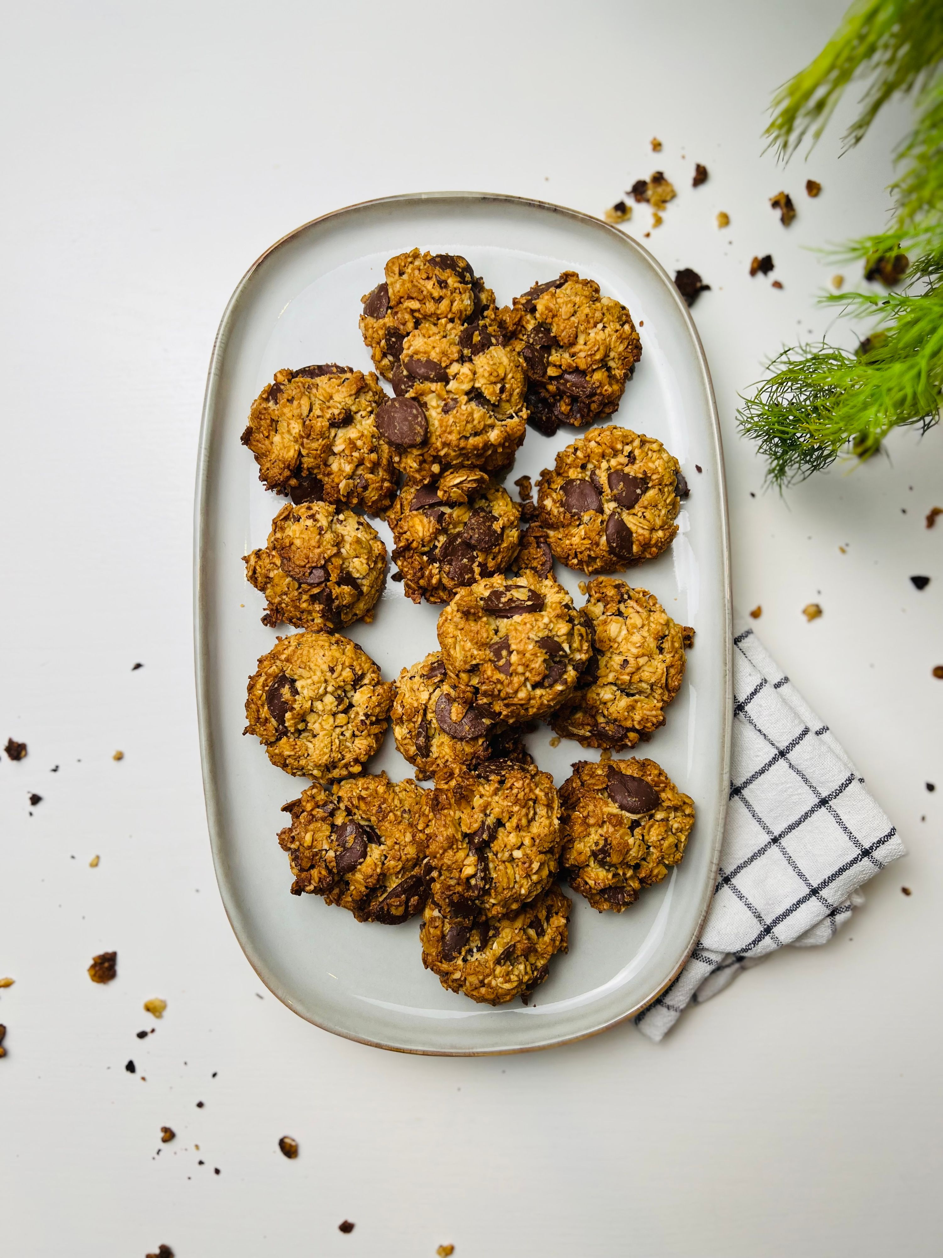 Overhead shot of a platter piled with crunchy chocolate chip oatmeal cookies on a shiny white countertop. A small branch of rosemary is in top right with crumbs scattered on the counter. A neatly folded white tea towel with thin gray lines peeks out from under the platter.