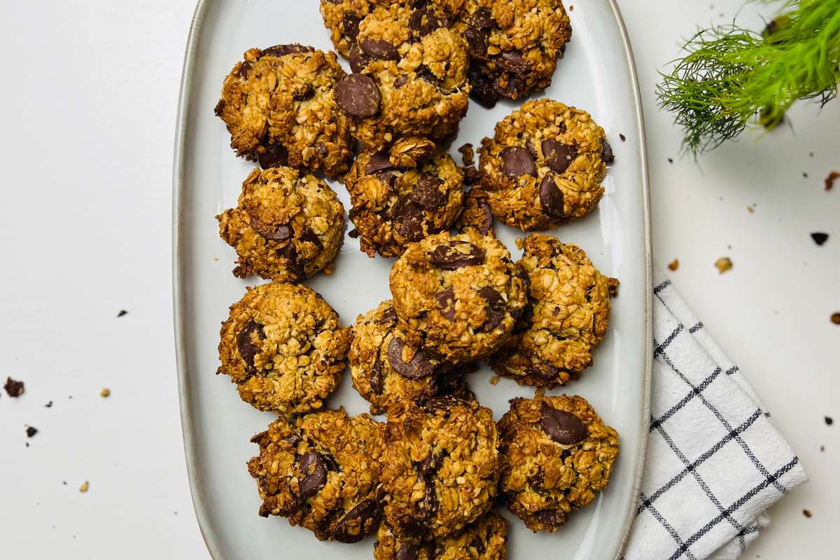 Overhead shot of a platter piled with crunchy chocolate chip oatmeal cookies on a shiny white countertop. A small branch of rosemary is in top right with crumbs scattered on the counter. A neatly folded white tea towel with thin gray lines peeks out from under the platter.