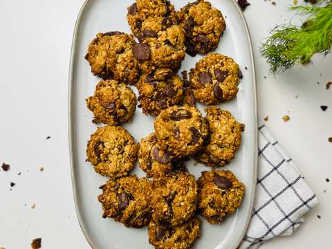 Overhead shot of a platter piled with crunchy chocolate chip oatmeal cookies on a shiny white countertop. A small branch of rosemary is in top right with crumbs scattered on the counter. A neatly folded white tea towel with thin gray lines peeks out from under the platter.