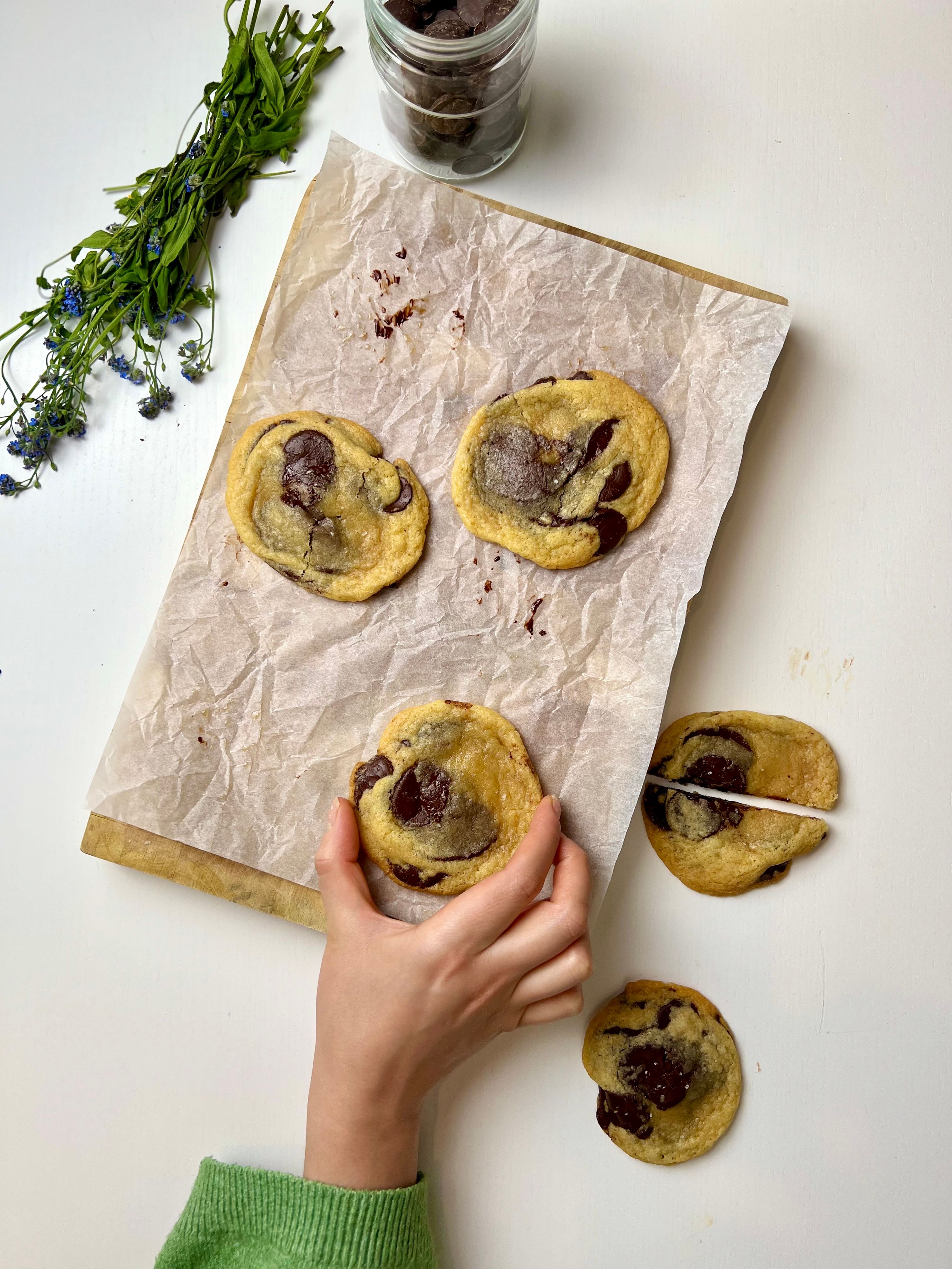Close-up of a golden-brown chocolate chip cookie with sea salt on top, resting on a cooling rack, with a glass of milk in the background.