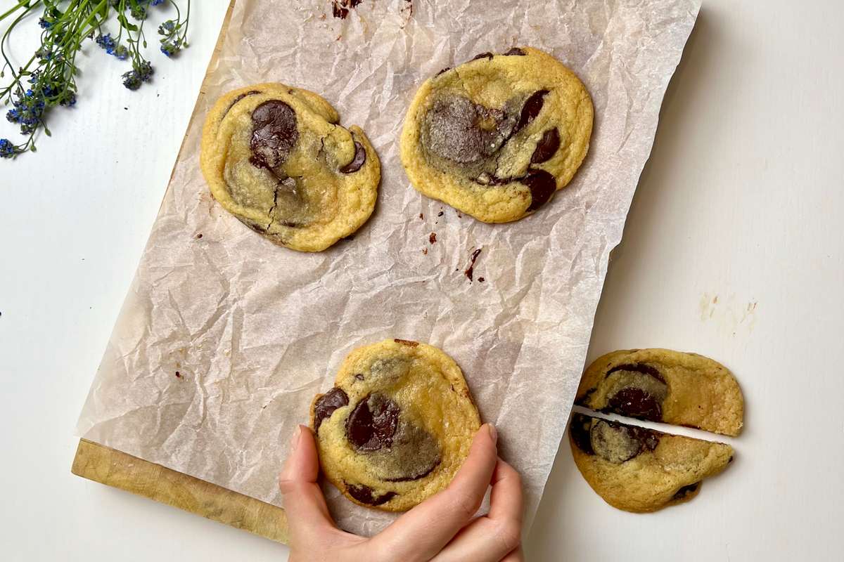 Close-up of a golden-brown chocolate chip cookie with sea salt on top, resting on a cooling rack, with a glass of milk in the background.
