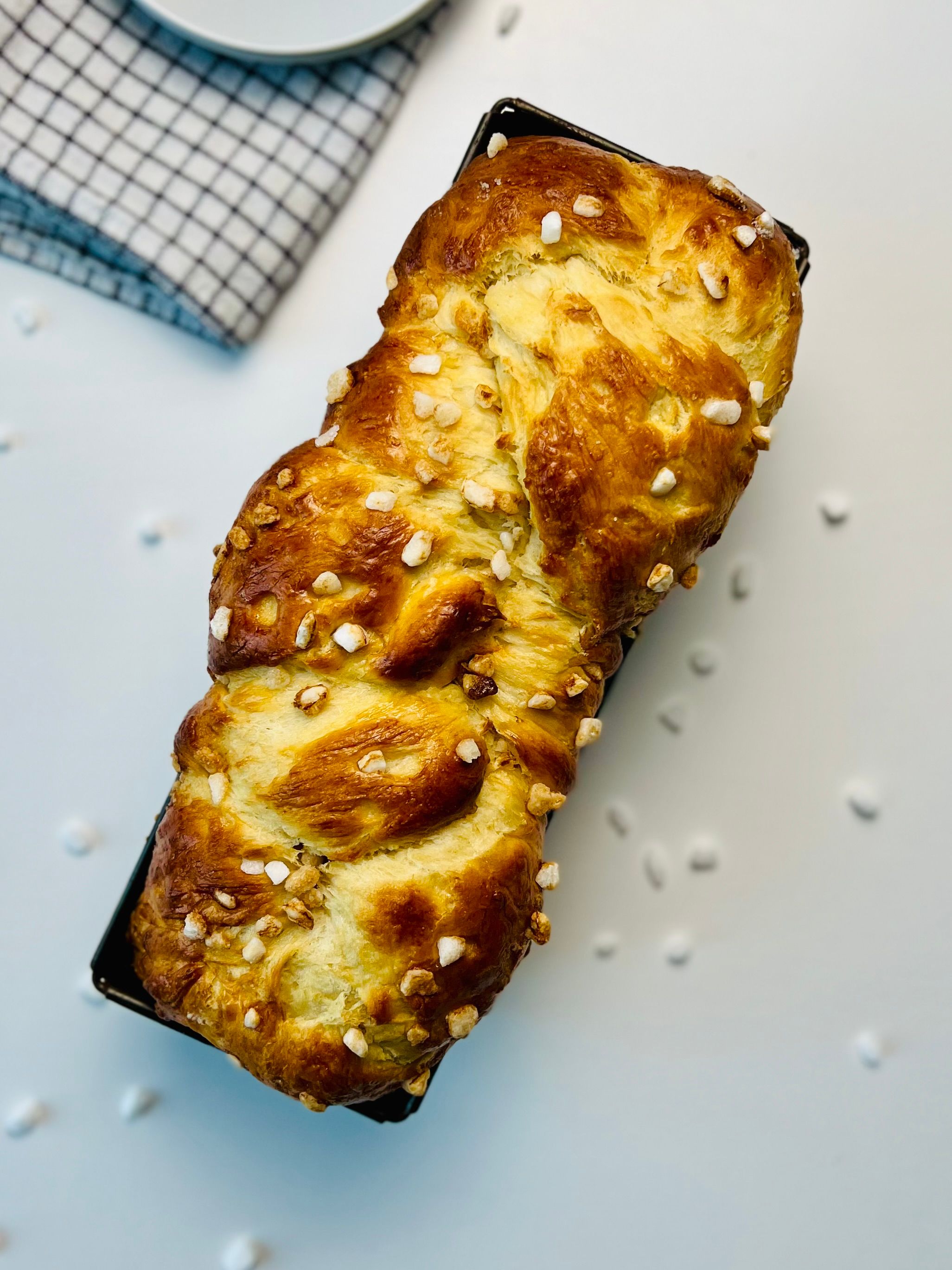 Overhead view of a brioche loaf, golden brown and braided, topped with pearl sugar, resting in a black loaf pan on white surface with a checkered napkin in the background.