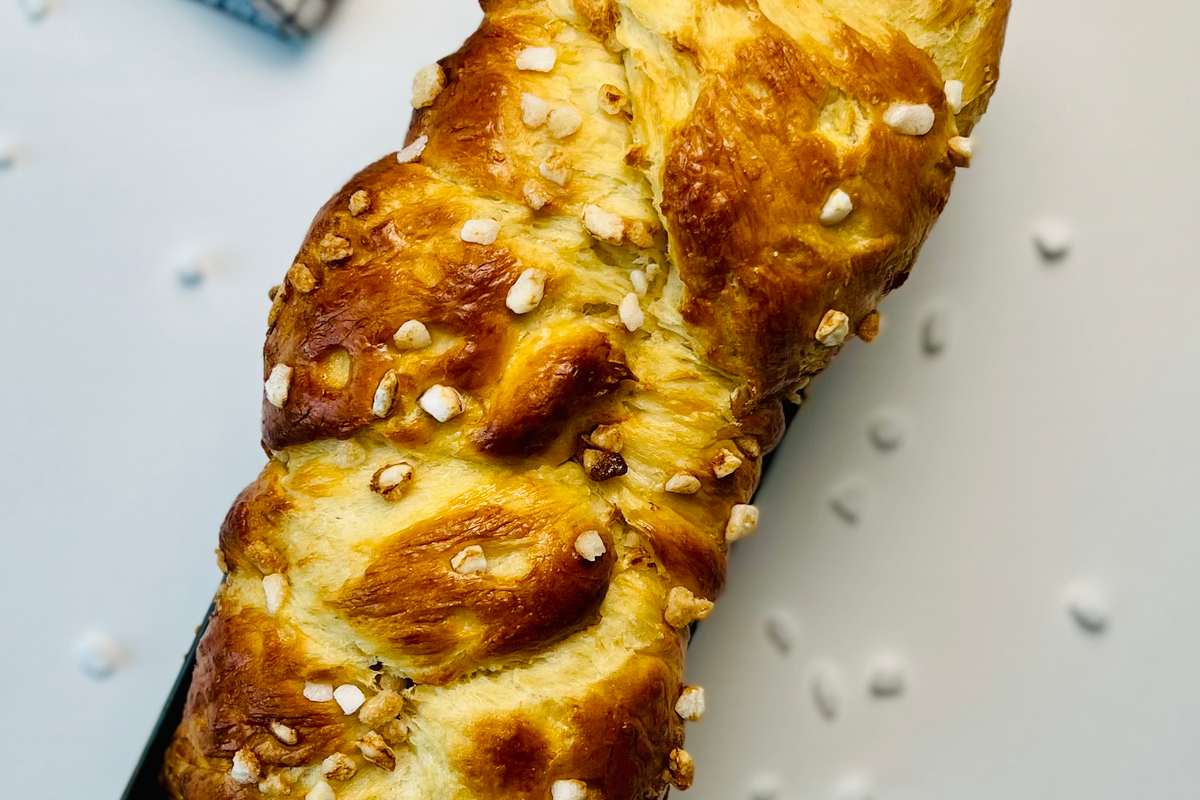 Overhead view of a brioche loaf, golden brown and braided, topped with pearl sugar, resting in a black loaf pan on white surface with a checkered napkin in the background.