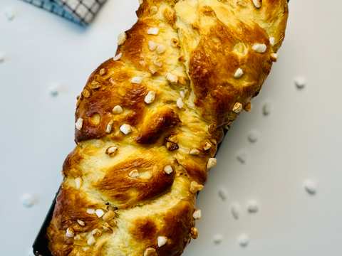 Overhead view of a brioche loaf, golden brown and braided, topped with pearl sugar, resting in a black loaf pan on white surface with a checkered napkin in the background.