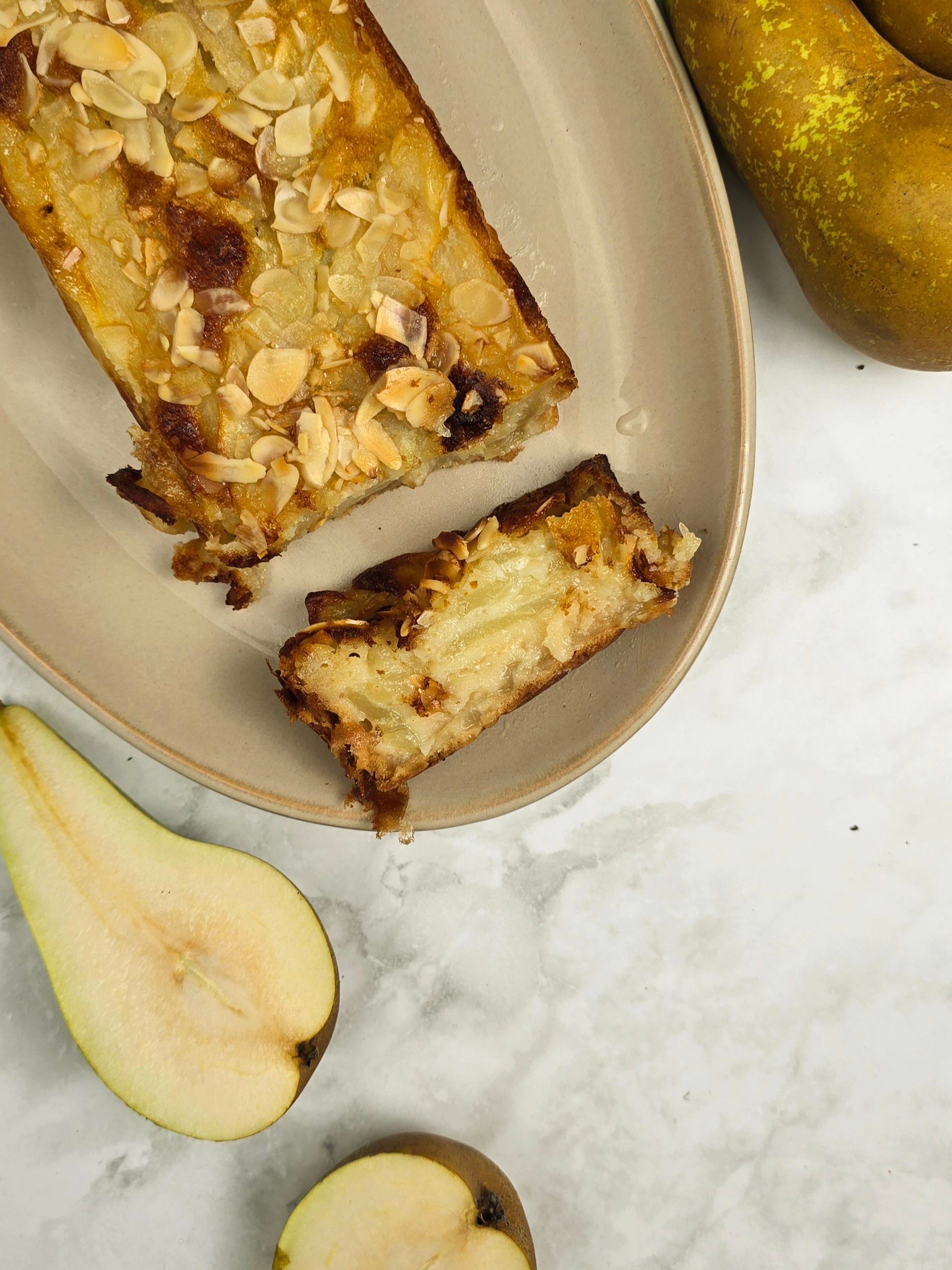 Close-up of a pear and almond cake slice on a beige plate. The cake is golden brown with toasted almond flakes on top.