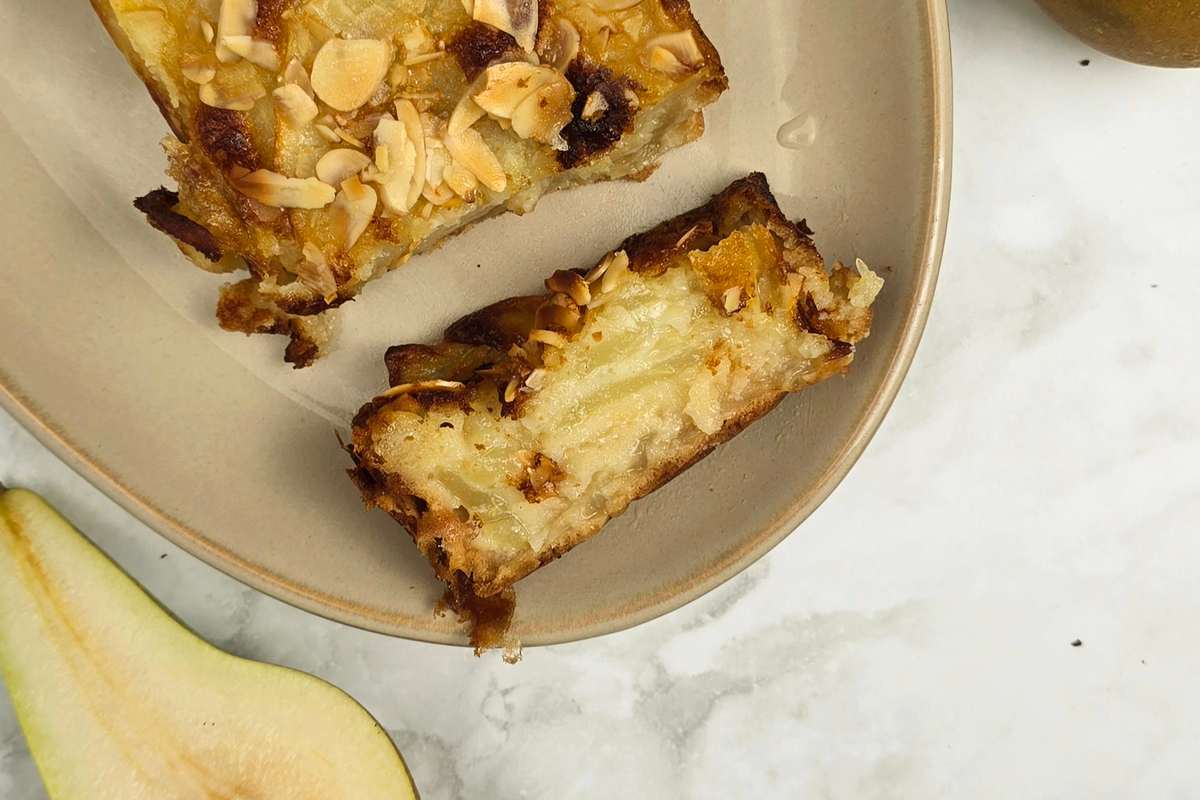 Close-up of a pear and almond cake slice on a beige plate. The cake is golden brown with toasted almond flakes on top.