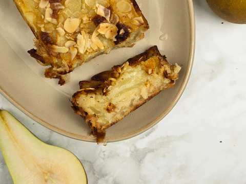 Close-up of a pear and almond cake slice on a beige plate. The cake is golden brown with toasted almond flakes on top.