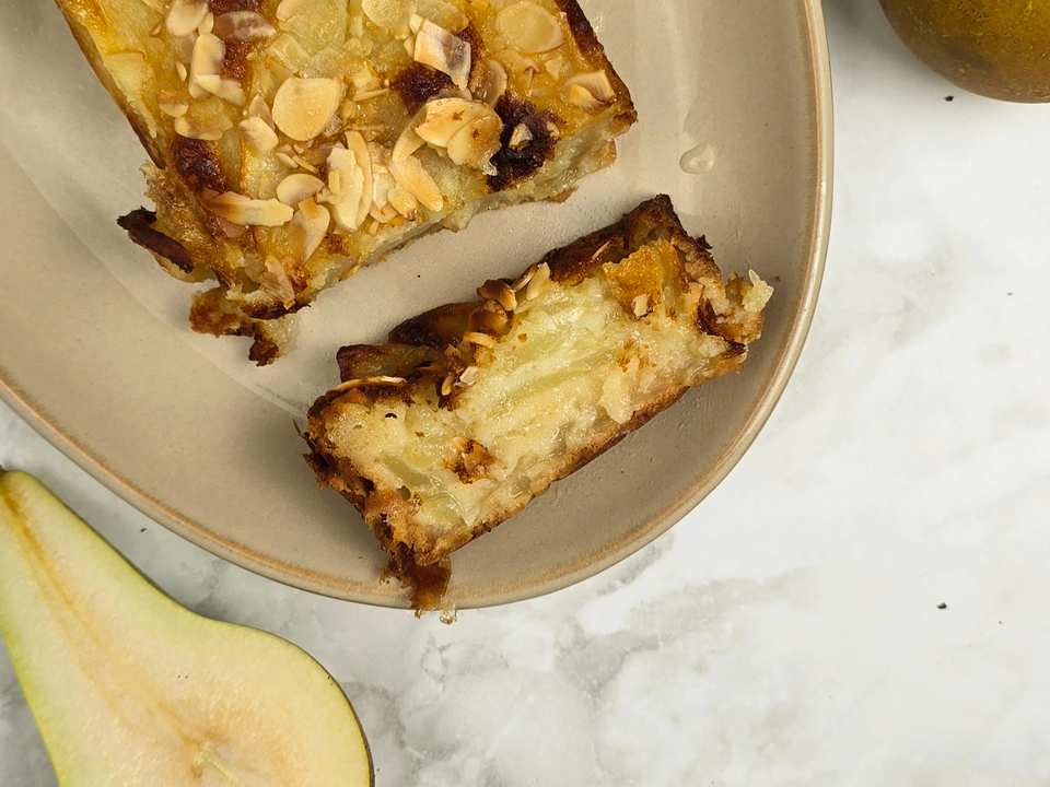 Close-up of a pear and almond cake slice on a beige plate. The cake is golden brown with toasted almond flakes on top.